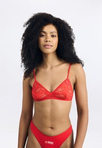 Bralette en dentelle rouge avec de fines bretelles et une ceinture assortie. Présente des motifs floraux et un petit logo sur la ceinture. Texture lisse.