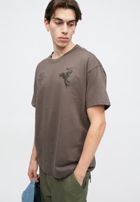 Καφέ βαμβακερό t-shirt με χαλαρή εφαρμογή, με μαύρη γραφική εκτύπωση στο στήθος και στρογγυλή λαιμόκοψη. Συνδυάζεται με ελιόχρωμα παντελόνια.