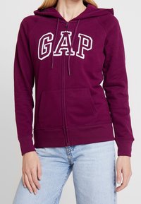 Femme portant un sweat à capuche zippé violet avec le logo "GAP" en lettres blanches et un jean bleu clair, vue du cou aux hanches.