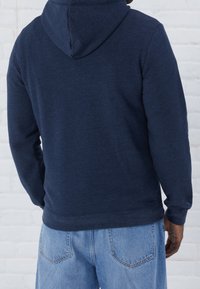 Marinblå hoodie tillverkad av texturerat tyg, med en bakvy som visar huva och långa ärmar, i kombination med ljusblå denimjeans.