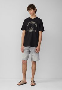 Zwarte katoenen T-shirt met een bedrukte graphic in beige, gecombineerd met lichte denim shorts en bruine sandalen met twee banden en een gesp.
