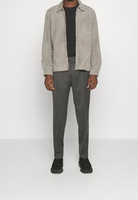 Szara zamszowa kurtka na suwak na czarnej koszuli, w połączeniu z szarymi spodniami w paski i czarnymi sneakersami. Kurtka posiada kieszeń na piersi.