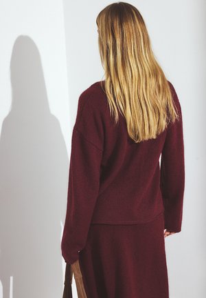 Bordeaux gebreide trui met een relaxed fit, verlaagde schouders en geribde zoom, gecombineerd met een bijpassende rok. De textuur lijkt zacht en comfortabel te zijn.