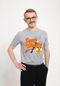 Grå t-shirt med korte ærmer, der har farverig "Izzy Wizzy" tekst, en tryllestav grafik, og en illustration af et plysdyr. Bæret med sorte bukser og et bælte.