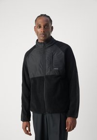 Svart jakke med glidelås foran, laget av fleece og nylon. Har en mock krage, sidelommer og en brystlomme med logodetalj.
