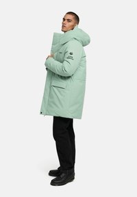 Jasnozielona puchowa kurtka z wysokim kołnierzem, przednimi kieszeniami i logo na rękawie. Połączona z czarnymi spodniami i botkami.