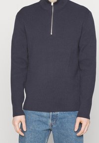 Granatowy sweter z prążkowanego dzianiny z wysokim kołnierzem i półzamkiem z przodu. Długie rękawy, dopasowany krój i fakturowana tkanina. Połączony z niebieskimi dżinsami.