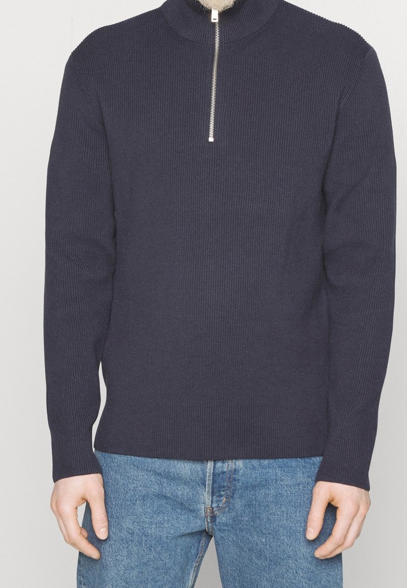 Granatowy sweter z prążkowanego dzianiny z wysokim kołnierzem i półzamkiem z przodu. Długie rękawy, dopasowany krój i fakturowana tkanina. Połączony z niebieskimi dżinsami.
