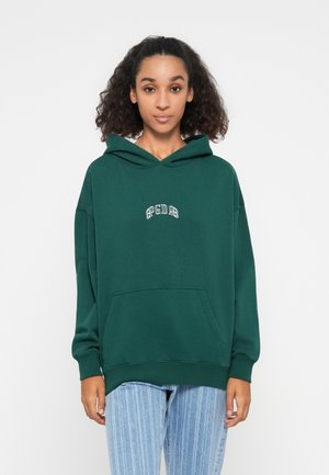 Green hooded sweatshirt with a front pocket, featuring white embroidered text. Worn with light blue vertically striped pants.
