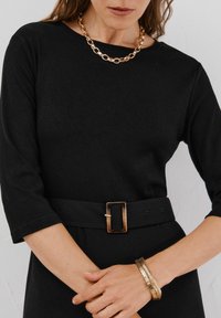 Czarny top z rękawami trzy czwarte, taliowany w pasie z czarnym paskiem z prostokątną brązową klamrą oraz złotym łańcuchem na szyi.