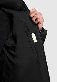 Chaqueta negra aislante con abertura con cremallera, que presenta un interior de tela y una pequeña etiqueta en uno de los bolsillos interiores.