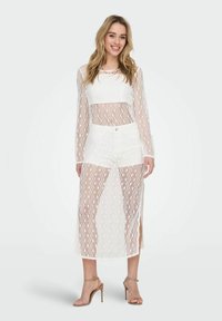 Weißes Spitzenkleid mit langen Ärmeln und einer Länge bis zur Mitte der Wade. Verfügt über geometrische Muster und einen hohen Schlitz. Kombiniert mit beigen Keilsandalen.