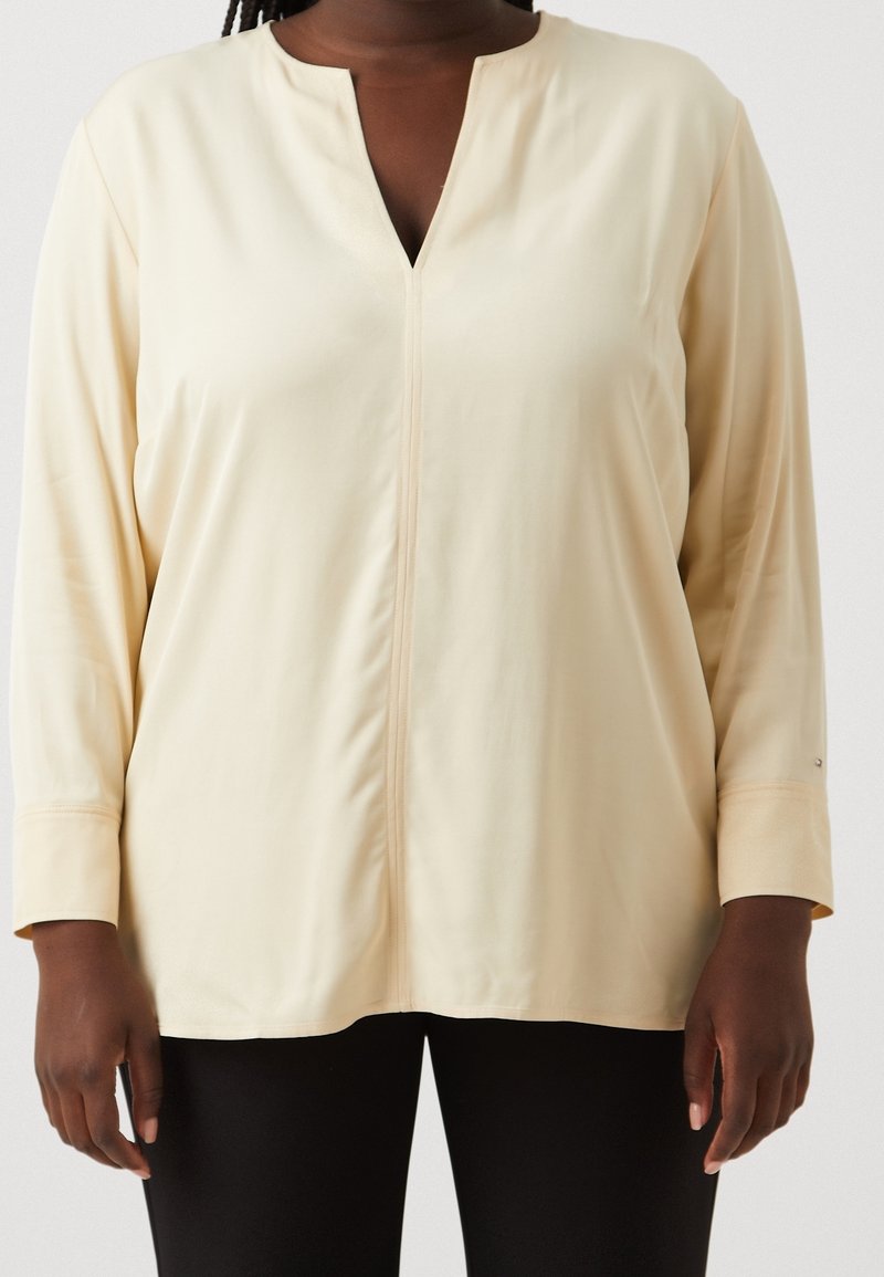 Haut à manches longues beige clair avec un col en V et une couture frontale. Tissu doux et lisse avec une coupe décontractée et des manches retroussées.