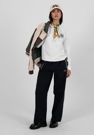 Frau trägt ein weißes Sweatshirt, schwarze Hose, eine beige Mütze und ein gemustertes Halstuch, hält eine Jacke über der Schulter.