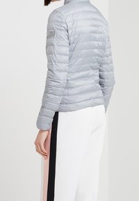 Ljusgrå pufferjacka med horisontell kviltning, hög krage och logotypdetalj på ärmen. Kombinerad med vita byxor med färgade ränder.