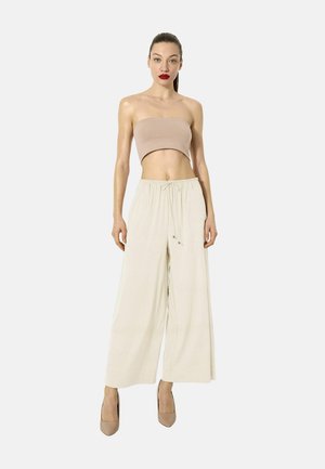 Femme aux cheveux plaqués en arrière, portant un top beige tube, un pantalon large crème avec cordon de serrage et des talons hauts beiges, debout devant un fond uni.