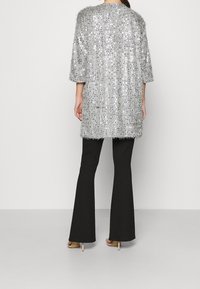 Silver, paljettbeströdd cardigan med en lös, texturerad design och trekvartslånga ärmar, i kombination med svarta utsvängda byxor och nude-häckar.