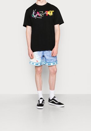 Persona vistiendo una camiseta negra con gráficos y texto colorido, pantalones cortos azul claro con estampado, calcetines blancos y zapatillas negras, de pie contra un fondo liso.