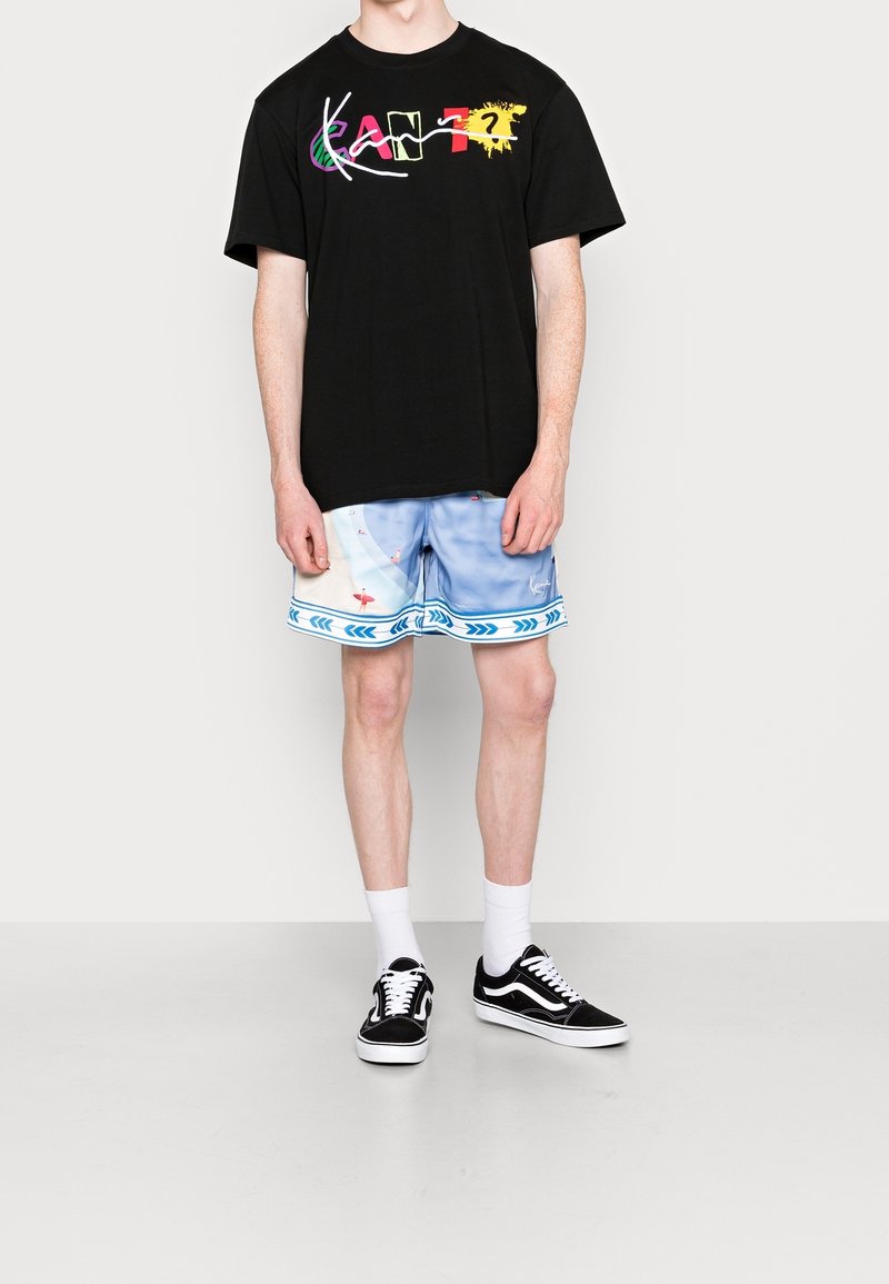 Personne portant un t-shirt graphique noir avec un texte coloré, un short bleu clair à motifs, des chaussettes blanches et des baskets noires, debout devant un fond uni.