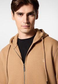 Kapturowa bluza z zamkiem w kolorze jasnobeżowym, wykonana z miękkiego materiału. Posiada czarny zamek oraz subtelną grafikę na piersi, noszona na czarną koszulkę.