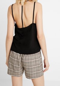 Top nero senza maniche con spalline sottili e schiena bassa, abbinato a pantaloni corti a quadri marrone chiaro. Il materiale sembra liscio con una vestibilità rilassata.