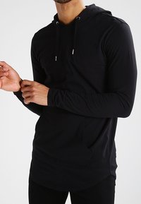 Černá mikina s dlouhým rukávem, klokaní kapsou, kapucí se stahovací šňůrkou a slim fit střihem. Tkanina vypadá měkce s hladkým povrchem.