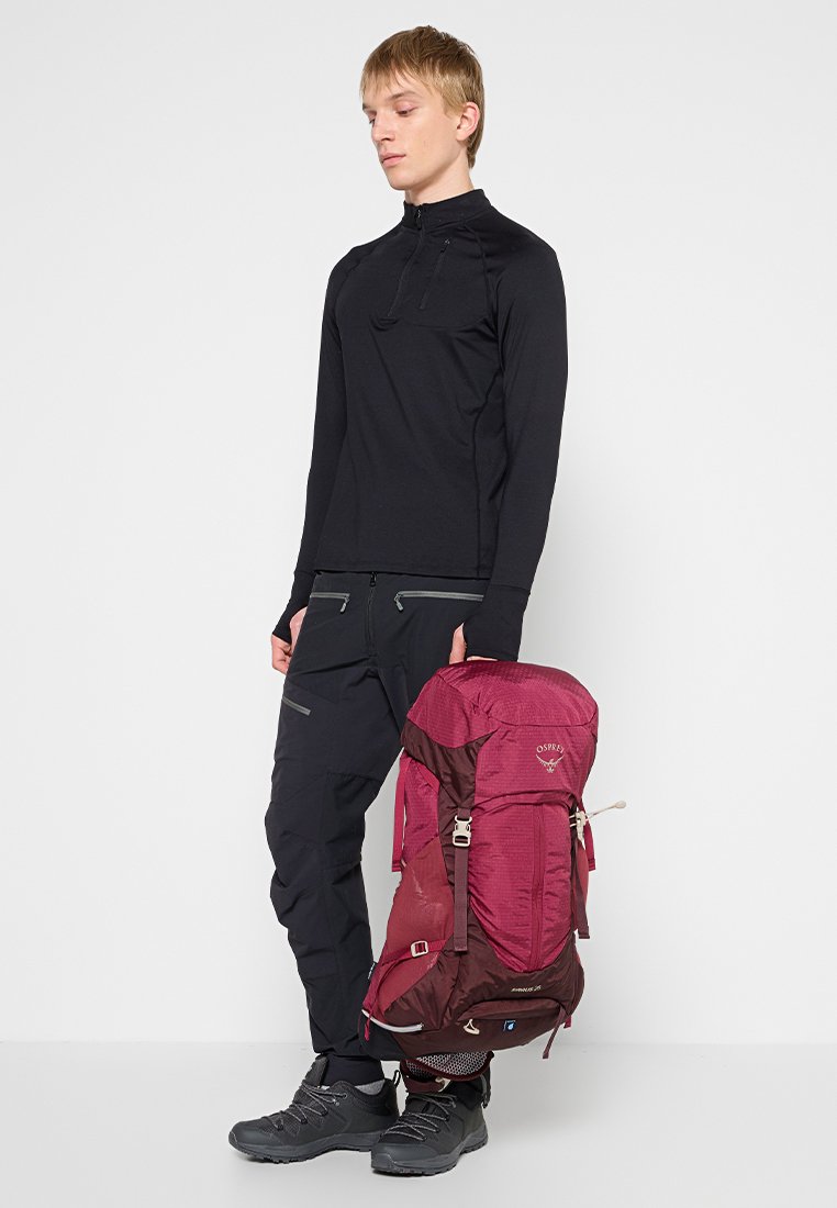 Jonge man in zwarte buitensportkleding met een grote rode wandeltas, staande tegen een effen lichte achtergrond.