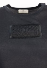 Camiseta de algodón negra con un parche de tejido negro texturizado y logotipo en relieve en el pecho, cuello redondo y mangas cortas.