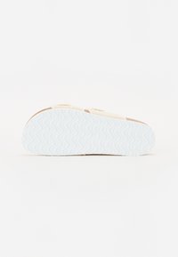 Fottøy med en teksturert hvit gummisåle med et bølgete mønster og en overdel i beige semsket skinn. To justerbare borrelåser er synlige.