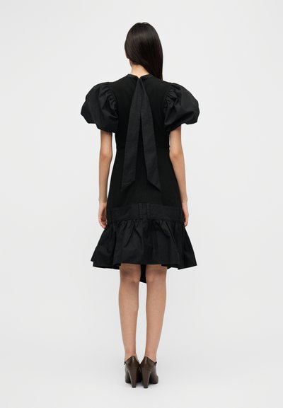 Μαύρο φόρεμα με φουσκωτά μανίκια, λεπτομέρεια με δέσιμο στην πλάτη και βολάν στο τελείωμα. Λείο ύφασμα και στενή κορυφή, μήκος μέχρι το γόνατο. Καφέ παπούτσια με τακούνι.