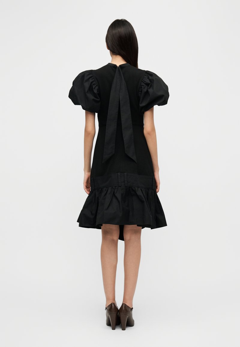 Abito nero con maniche a sbuffo, dettaglio annodato sul retro e orlo arricciato. Tessuto liscio e corpetto aderente, lunghezza al ginocchio. Scarpe con tacco marroni.