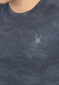 Mørkeblå sportsbluse med et tekstureret camouflage mønster, der har et diskret edderkop-logo i midten. Glat stof med rund halsudskæring.
