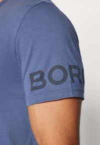 Mėlyna trumpa rankovė marškinėliai su tekstūruotu audiniu. Kairėje rankovėje buvo didelis juodas užrašas "BORO".