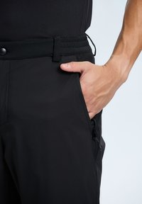 Zwarte sportbroek met elastische tailleband, zijzak met ritssluiting, gladde textuur en gestroomlijnd ontwerp, geschikt voor actief gebruik.