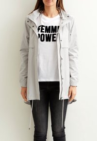 Lysegrå jakke med lange ærmer og hætte, frontlynlås og lommer. Worn over en hvid t-shirt med sort tekst, kombineret med sorte jeans.