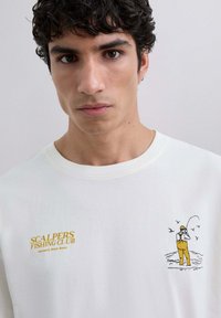 Tânăr cu părul curly închis la culoare, purtând un hanorac alb cu text galben "Scalpers Fishing Club" și o ilustrație a unui pescar.