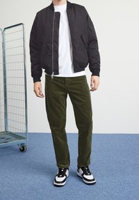 Svart bomberjacka med dragkedja, vit T-shirt under, olivgröna kordflortbyxor och svarta sneakers med vita accenter och snören.