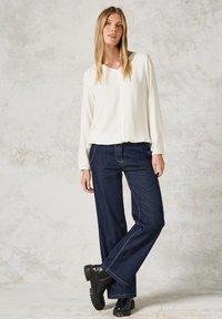 Vit långärmad blus med v-ringning och texturerad tyg, i kombination med mörkblå vida jeans och kraftiga svarta stövlar.