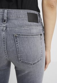 Jeans de mezclilla gris de cintura alta, con un parche de cuero negro, costuras visibles y bolsillos traseros con detalles metálicos.