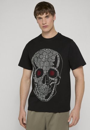 Jeune homme portant un t-shirt noir avec un grand motif de crâne blanc à pois avec des yeux rouges, debout devant un fond neutre.