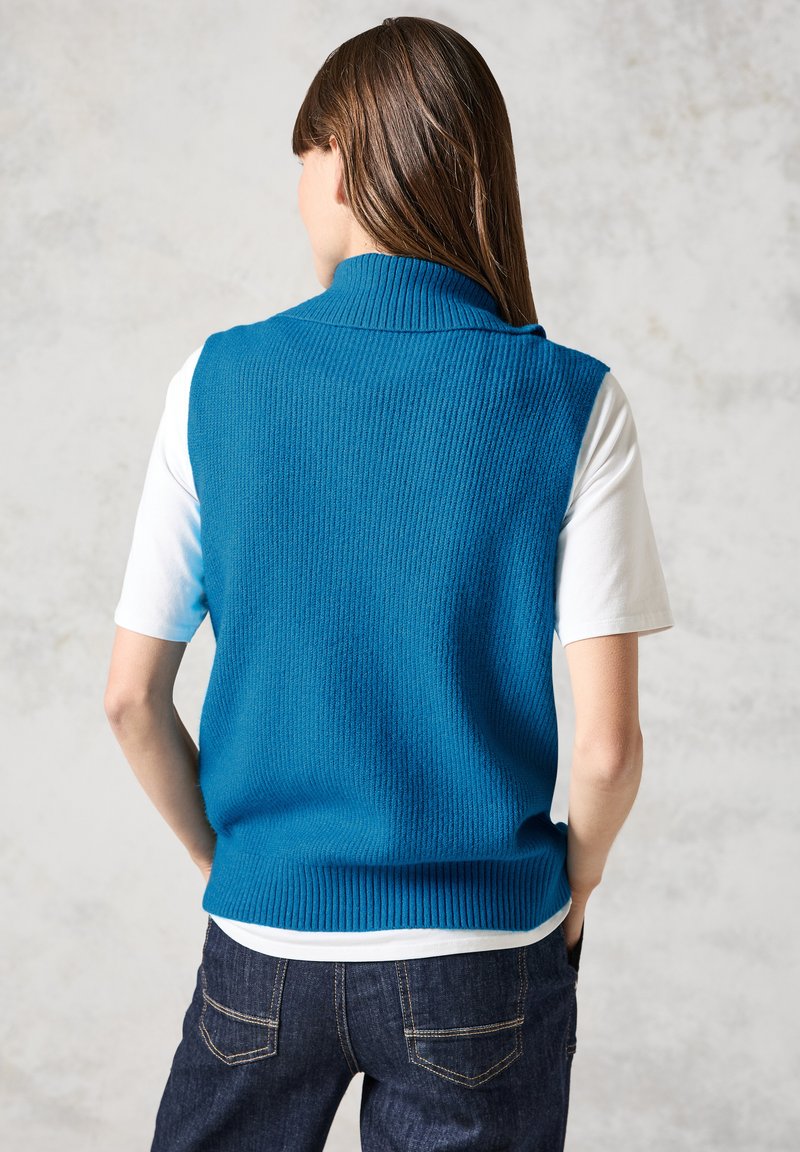 Bezramenná modrá pletená vesta s vysokým límcem, nošená přes bílý tričko s krátkým rukávem, v kombinaci s tmavě modrými džíny.