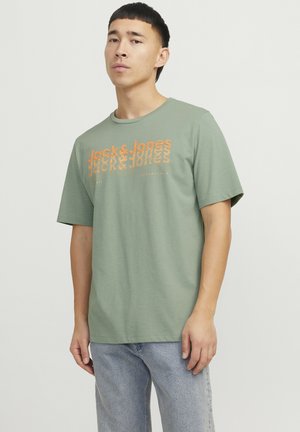 Man met kort donker haar, gekleed in een saliegroene Jack & Jones t-shirt en lichtblauwe jeans, staand tegen een effen lichte achtergrond.