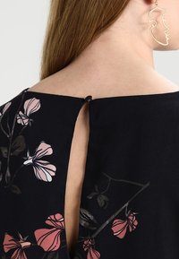 Blusa negra con estampados florales en tonos rosa y crema, con una pequeña abertura en forma de llave en la parte posterior y una textura de tela suave.
