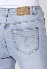 Jean en denim bleu clair avec poche arrière présentant une couture courbée et une étiquette de marque en cuir marron indiquant "Koroshi Denim Collection."
