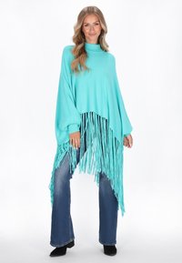 Naine seisab seljas teal-värvi fransiõlgpikkuse poncho, sinised laiad teksad ja mustad kontsaga saapad valge taustaga.