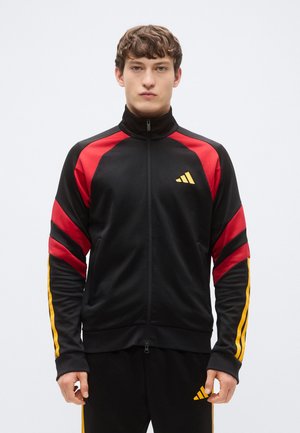 Μαύρο αθλητικό μπουφάν με κόκκινες λεπτομέρειες, κίτρινες ρίγες στα μανίκια, ψηλό γιακά και φερμουάρ μπροστά· διαθέτει το λογότυπο της Adidas σε κίτρινο.