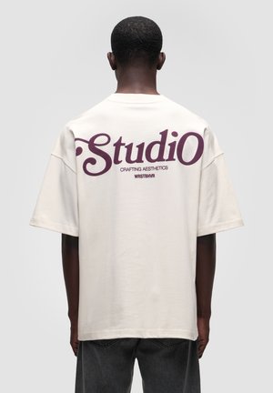 Man draagt een losse witte T-shirt met de tekst "StudiO Crafting Aesthetics WRSTBHVR" in paars op de rug, staand tegen een effen achtergrond.