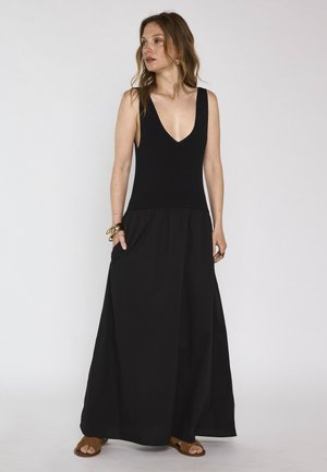 Femme portant une robe longue noire sans manches avec un décolleté en V profond et des sandales marron, debout avec une main dans la poche.