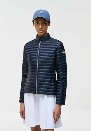 Donna con giacca trapuntata blu con cerniera frontale, gonna bianca e cappellino azzurro chiaro, in piedi davanti a uno sfondo chiaro uniforme.