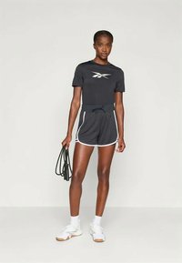 T-shirt atlético preto com logótipo branco, combinado com calções escuros com acabamento branco. O modelo segura uma corda de saltar, vestindo sapatilhas atléticas brancas.